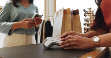Kadın, butik mağazası müşterisi. Telefonunu NFC teknolojisiyle kullanıyor. Satıcı kadın da kart okuyucu makinesini tutuyor. İşlem, ulaşılamaz yöntem ödeme, alışveriş