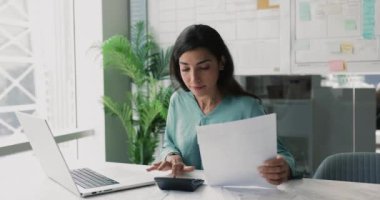Dizüstü bilgisayarda çalışan ve faturaları ödemek için hesap makinesi kullanan muhasebeci kadın, gülümseme, faturaları ve faturaları gözden geçirme, bütçeyi izleme, gelir ve giderleri izleme, yıllık mali rapor hazırlama