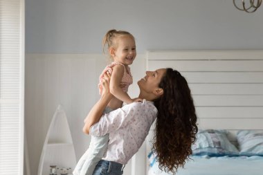 Happy embraces. Affectionate millennial mom lift little preschooler daughter kid up in air for loving hug bonding single parent and child cuddling laughing together at warm cozy bedroom in family home clipart