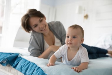 Çocuk gelişimi ve güvenliği. Gülümseyen genç anne dadı büyük rahat bir yatakta uzanıp emeklemeyi öğrenen bebek bebeği seyrediyor. Dördünün de üzerinde duran, bilişsel ilgiyle ileriye bakan bebek çocuğa odaklan.
