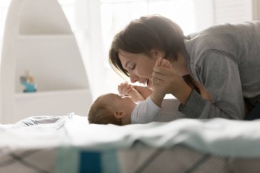 Ebeveyn eğlencesi. Gülen genç bir anne, küçük şirin bir çocukla evde vakit geçirmekten büyük yatakta yatıp bebekler için jimnastik yaparken eğlenmekten zevk alıyor. Boşluğu kopyala