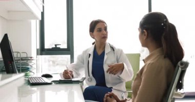 Ciddi Orta Doğulu kadın doktor hasta kadınla sağlık şikayetleri hakkında konuşuyor, çalışma masasında oturuyor, hastane ofisinde danışmanlık yapıyor, notlar yazıyor, tıbbi kayıtları tutuyor.