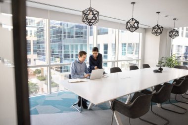 Ofis toplantı odasında dizüstü bilgisayardan online içeriği izleyen, büyük konferans masasında tek başına oturan, online iletişimi takım çalışması için kullanan çok ırklı iki pozitif yönetici. Geniş açılı çekim