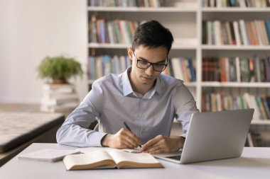 Ev ödevi. Üniversitenin kütüphanesinde edebiyat eleştirisi yazan gözlüklü genç bir erkek öğrenci bilim yarışması için araştırma projesi üzerinde çalışıyor. Not defterindeki kağıt kaynakları inceliyor.