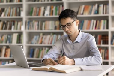 Öğrenmekle meşgul. Kütüphane masasında gözlük takan ciddi motivasyona sahip genç bir erkek öğrenci ödevlerini deftere yazar not alır bilgiyi çevrimiçi edinmek için not defteri kullanır. Boşluğu kopyala