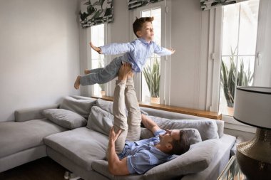 Sevimli çocuk ve güçlü baba kanepede uçak uçuruyor. Baba sırt üstü uzanıyor, çocuğa destek oluyor, kollarını açıyor, akroyoga yapıyor, oğluyla birlikte egzersiz yapıyor.