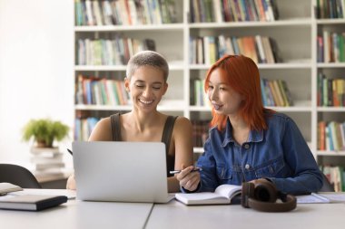 Uzaktan öğrenme. İki sırıtkan genç kız üniversite öğrencileri kütüphanede çevrimiçi eğitim için dizüstü bilgisayar kullanıyor. Video dersi alın ve web sitelerinde sörf hakkında bilgi hazırlayın.