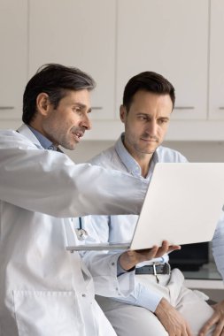 Ciddi orta yaşlı pratisyen doktor hastaya tıbbi rapor gösteriyor, dizüstü bilgisayara işaret ediyor, test sonuçlarını açıklıyor, modern teknolojiyi danışmanlık için kullanıyor.