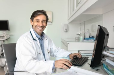 Neşeli, orta yaşlı, bilgisayar başında çalışan Latin doktor. Kameraya bakıyor, klinik çalışma masasında profesyonel tıbbi portre için poz veriyor, gülümsüyor, gülüyor.