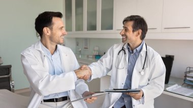 Klinik muayene odasında tokalaşan iki mutlu erkek doktor. Pozitif latin doktorunun iş arkadaşına el sıkışması takım çalışması, danışmanlık ve başarılı bir işbirliği için teşekkür etmesi. Sancak vuruşu