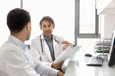 Orta yaşlı İspanyol doktor ofisteki iş yerinde hastayla konuşuyor, kağıt sağlık raporunu açıklıyor, konsültasyon yapıyor, tıbbi tavsiye veriyor, reçete yazıyor, tedavi detaylarını anlatıyor.