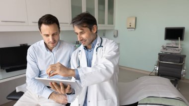 Odaklanmış orta yaşlı Latin doktor hastalara muayene raporları gösteriyor, ultrason tarama sonuçlarını açıklıyor, tablet ekranı işaret ediyor, hastalarla çalışmak için elektronik teknolojiyi kullanıyor.