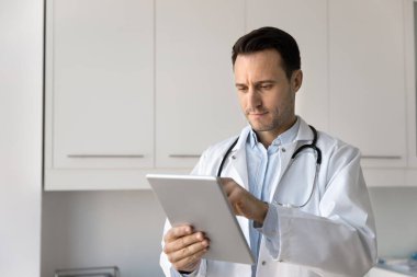 Ciddi kendine güvenen erkek pratisyen doktor çevrimiçi servisi dijital tablet üzerinde kullanıyor, elektronik tıp raporlarını okuyor, muayene kayıtları, sağlık araştırmaları sonuçları, internette geziniyor.