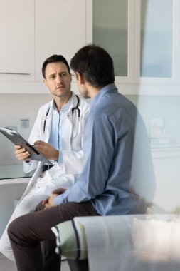 Odaklanmış doktor, erkek hastayı muayene ediyor, klinikte çalışıyor, not defterine notlar yazıyor, sağlık sorunlarından bahsediyor, kanepede oturuyor. Dikey gizli çekim