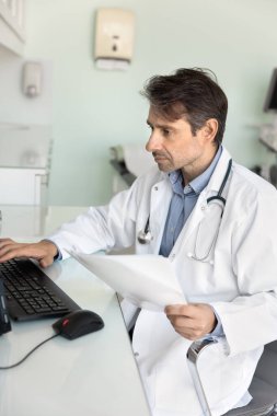 Masaüstünde çalışan orta yaşlı doktor doktor, kağıt dökümanlarını inceliyor, sınav sonuçlarını klavyeye yazıyor, elektronik tıp raporu yazıyor. Dikey çekim
