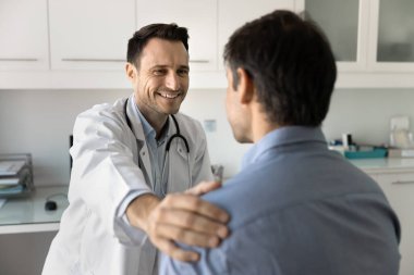 Neşeli erkek doktor hastanın omzuna dokunuyor, destekleyici dostça bir jest yapıyor, destek veriyor, sempati gösteriyor, iyi haberler veriyor, iyimser teşhis koyuyor, başarılı terapi sonuçları veriyor.