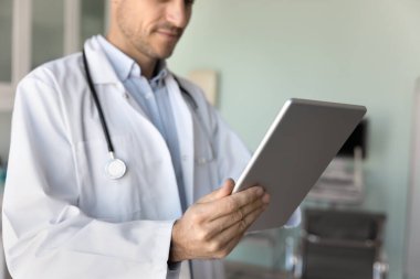 Modern teknolojiyi kullanan beyaz önlüklü erkek doktor, hastane ofisinde tabletle çalışıyor, hastaların geçmiş kayıtlarını okuyor, tıbbi muayene elektronik raporlarını okuyor. Kırpılmış atış