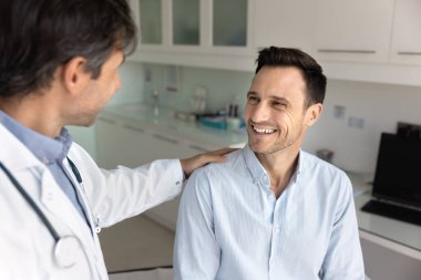 Neşeli sabırlı adam doktordan destekleyici omuz hareketi alıyor, muayene odasında kanepede oturuyor, sağlık uzmanını muayene ediyor, test sonuçları hakkında iyi haberler öğreniyor.