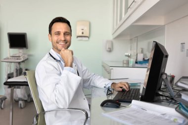 Ultrason tarama odasındaki bilgisayarın başında çalışan mutlu doktor, kameraya bakıyor, tıbbi profesyonel portre için poz veriyor, çeneye dokunuyor, gülümsüyor, masaüstü ile çalışma masasında oturuyor.