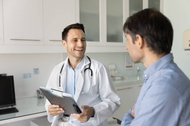 Neşeli iyimser doktor hastaya danışmanlık yapıyor, gülümseyen bir adamla konuşuyor, check-up 'tan sonra iyi haberler veriyor, tedaviyi, terapi detaylarını anlatıyor, kağıt belgeler tutuyor, kayıtlar tutuyor.