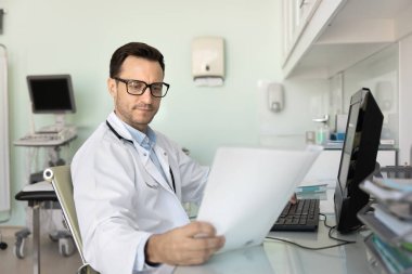 Ciddi genç doktor gözlüklü ve beyaz önlüklü. Masaüstü bilgisayar kullanarak iş yerinde kağıt tıp raporu okuyor. Sonograf ultrason taraması sonuçlarını analiz ediyor, belgeyi gözden geçiriyor,