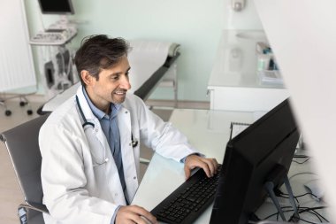 Hastane muayene odasındaki bilgisayarda çalışan, klavyeye yazı yazan elektronik kayıtları okuyan, gülümseyen, modern internet teknolojisini tıp alanında çalışan Latin bir doktor.