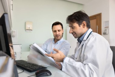 Pozitif orta yaşlı doktor hastaya danışmanlık yapıyor, kağıt geçmişi okuyor, tıbbi kayıtları analiz ediyor, teşhisi, tedaviyi, reçeteyi açıklıyor, dosyaya işaret ediyor, konuşuyor, gülümsüyor.