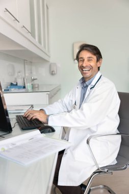 Hastane ofisindeki masaüstünde çalışan başarılı İspanyol pratisyen adam, kameraya bakıyor, gülümsüyor, klavyede yazıyor, çalışma masasında oturuyor. Dikey çekim portresi