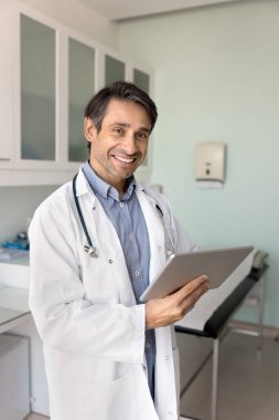 Neşeli İspanyol erkek doktor tablet üzerindeki elektronik tıbbi raporları analiz ediyor muayene odasında dijital cihaz kullanıyor, başka tarafa bakıyor, gülümsüyor, düşünüyor, modern teknolojiyle tıbbi işlerden keyif alıyor.