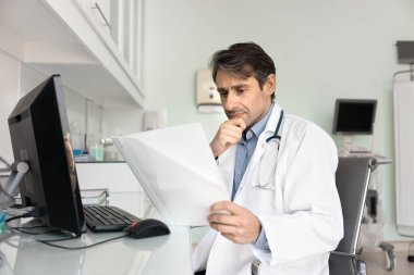 Odaklanmış Latin doktor belgeleri dikkatle okuyor, bilgisayarla işyerindeki tıbbi raporları inceliyor, çeneye dokunuyor, kayıtları, geçmişi, teşhisi, reçeteyi düşünüyor.