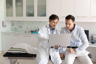 Ciddi orta yaşlı İspanyol doktor erkek hastaya elektronik tıbbi rapor gösteriyor, dizüstü bilgisayarını tutuyor, ekranı gösteriyor, muayene sonuçlarını anlatıyor, teşhis, tedavi