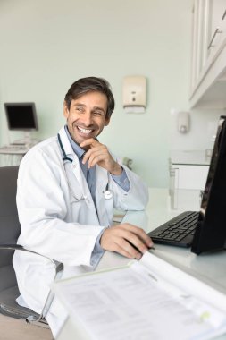 Başarılı orta yaşlı profesyonel doktor hastane ofisindeki çalışma masasında oturuyor, çenesine dokunuyor, başka tarafa bakıyor, gülümsüyor, gülüyor ve tıptaki internet teknolojisini düşünüyor.
