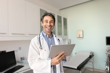 Tıbbi uzman portresi için poz veren orta yaşlı İspanyol doktor, dijital tablet tutuyor, muayene odasında duruyor, dişlek gülümsemeyle kameraya bakıyor, gülüyor.