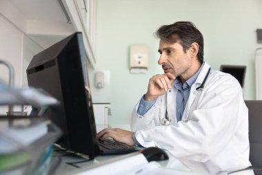 Odaklanmış orta yaşlı pratisyen doktor hastane ofisinde çalışıyor, test sonuçlarını okuyor, başvuru kayıtları, tıbbi raporlar, dokunaklı çene, düşünme, hastalarla çevrimiçi sohbet