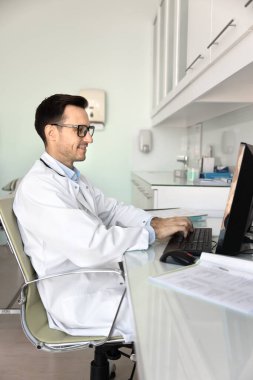 Bilgisayarda çalışan mutlu doktor, hastane ofisindeki çalışma masasında oturan, tıbbi rapor yazan, internette online danışmanlık yapan, profesyonel iletişim için masaüstünü kullanan.