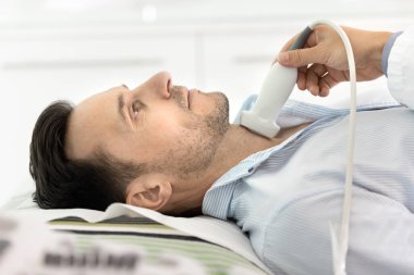 Tiroid muayenesi için sonografı ziyaret eden erkek hasta yan görüntüsü, kanepede uzanmış, boynunda tarayıcı var. Doktorun eli ultrason cihazı takıyor, boğaz hastalıklarını kontrol ediyor..