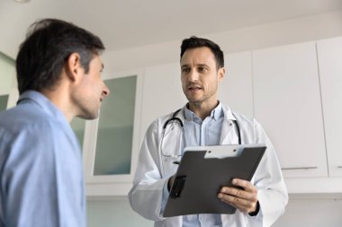 Ciddi bir pratisyen hekim, kardiyolog, klinikte hastayla konuşan, şikayetleri soran, kağıt kayıtlarına notlar yazan, danışmanlık yapan, tavsiye veren,