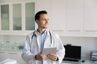 Tablet tutan, derin düşüncelere dalan, gülümseyen, tıptaki dijital teknolojiyi düşünen, hastaların elektronik kayıtlarını tutan, düşünceli genç bir doktor.