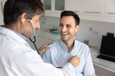 Orta yaşlı Latin pratisyen doktor mutlu hastanın göğsüne steteskop uyguluyor, kalp atışlarını kontrol ediyor, semptomları tartışıyor, şikayetleri tartışıyor. Neşeli adam kontrol için kardiyologları ziyaret ediyor.