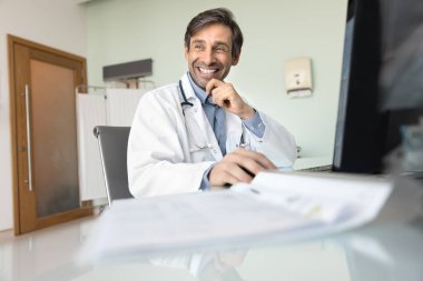Mutlu orta yaşlı pratisyen doktor hastane ofisinde bilgisayar başında çalışıyor, işyerinde oturuyor, dişlek bir gülümsemeyle başka tarafa bakıyor, başarılı bir iş davası üzerinde düşünüyor, internet teknolojisi.