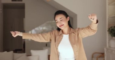 Mutlu genç 20 'li yaşlarda Asyalı bir kız, rahat bir dairede gözleri kapalı dans ediyor, eğleniyor, müzik dinliyor, rahatlıyor, gülümsüyor, başarıyı kutluyor, güzel bir konaklama ya da kiralama yapıyor.