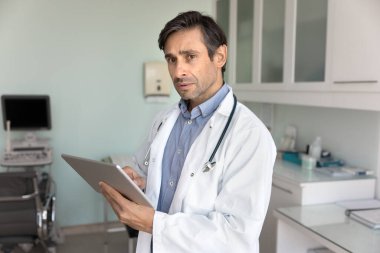 Ciddi bir Latin doktor, ultrason muayene odasında dijital tablet kullanıyor, başka yere bakıyor, hastaları analiz ediyor, sonuçları tarıyor, tıbbi rapor üzerinde düşünüyor, teşhis koyuyor.