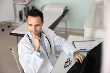 Klinik muayene odasında, iş yerindeki bilgisayarın başında oturan, çeneye dokunan, başka yere bakan tıptaki kariyerini düşünen, sağlık hizmetleri teknolojisini düşünen yakışıklı genç bir doktor.