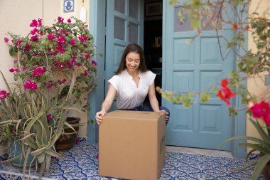 Beklenen paketi kapısından alan, lojistik ulaşım hizmeti tarafından teslim edilen büyük karton kutuya dokunan ve seçen mutlu genç müşteri kadın, gülümseyerek paketleri açmaya hazır.