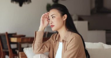 Depresyondaki genç Asyalı kadın kafasına dokunuyor ve kafasını çeviriyor, koltuğunda oturuyor, ciddi sorunları, sorunları, zor kararları, kötü haberleri, umutsuzca gözlerini kapatıyor.