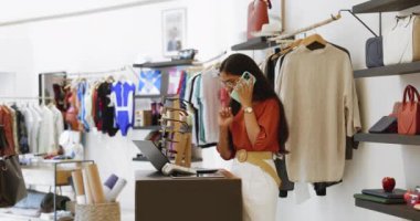 Modern butik mağazası sahibi veya satıcı dizüstü bilgisayarda çalışır, telefonda konuşur, nakit masasında durur, müşteri soruşturmasına cevap verir, tedarikçiyle siparişleri tartışır, operasyonel veya idari görevleri yönetir