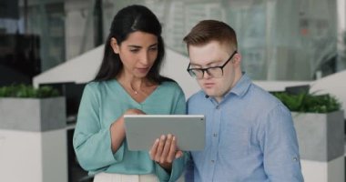 Down sendromlu genç adama görevi ya da yeni platformu açıklayan yetenekli genç bir kadın, iş günü birlikte dijital tablet kullanarak iş birliği yapıyor, öğrenme ya da takım çalışması yapıyor.