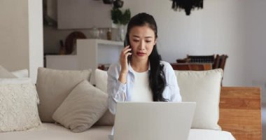 Odaklanmış Asyalı serbest çalışan kadın evde çalışıyor, iletişim için aletler kullanıyor, koltukta oturuyor, cep telefonuyla konuşuyor, dizüstü bilgisayarında telefon hizmeti veriyor, gülümsüyor.