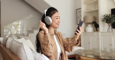 Kablosuz kulaklıklı neşeli Vietnamlı kız AI tarafından toplanan akıllı çevrimiçi müzik listesinden favori şarkıları dinliyor, çalışan medya uygulaması ile cep telefonu tutuyor, oturuyor, dans ediyor, gülümsüyor.