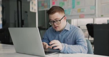 Down sendromlu ciddi bir adam modern şirket ofisinde dizüstü bilgisayarla çalışıyor çalışma masasında oturuyor, idari görevler öğreniyor, verileri gözden geçiriyor, çevrimiçi görevler yapıyor.